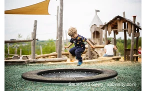 Junge springt auf einem Trampolin, im Hintergrund ein Spielplatz