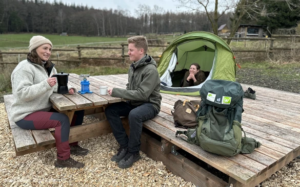 Zwei Wanderer sitzen an einem Tisch an einem Trekkingplatz im Arsnberger Wald. Im Hintergund steht ein Zelt, aus dem eine Frau schaut.
