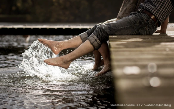 Zwei Ausflügler tauchen ihre Füße ins klare Wasser eines Sauerland-Sees.