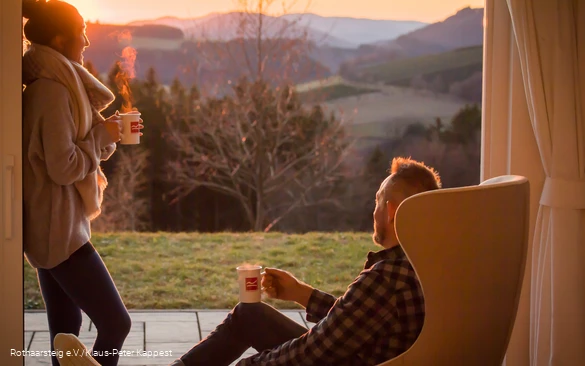 Ein Mann und eine Frau genießen mit einem heißen Getränk in der Hand ihren Blick aus dem Terrassenfenster auf die Sauerländer Landschaft